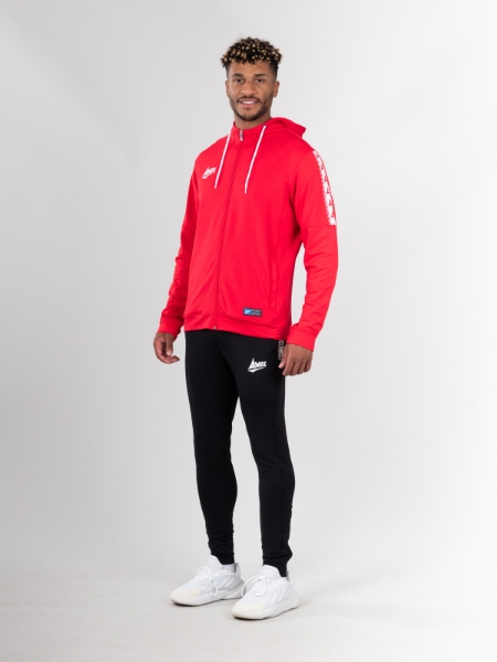 Person standing against a plain background, wearing a red zip-up hoodie with white drawstrings and a white logo on the left chest area. The hoodie features a white strip with red text along the left sleeve. The person is also dressed in black athletic pants with a white logo on the upper left thigh and white athletic shoes.