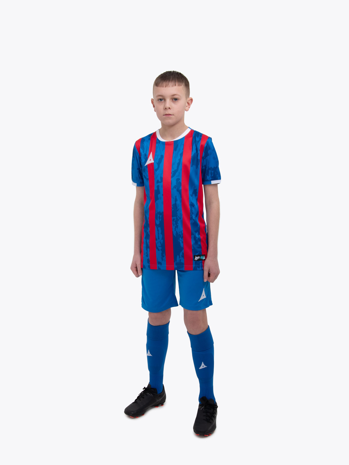 a kid standing up wearing a red and royal blue football shirt and matching socks and shorts 